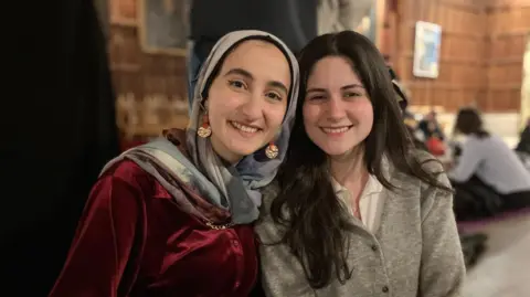 Raza, who is wearing a patterned headscarf and a red-velvet top, sits next to Maya, who is wearing a greyish cardigan. They are looking at the camera and smiling.