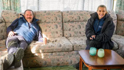 The image shows two people sitting on a beige sofa with floral patterns sewed on it. There is a brown coffee table with two candles on it. Behind the couch is a window covered with white lace curtains.