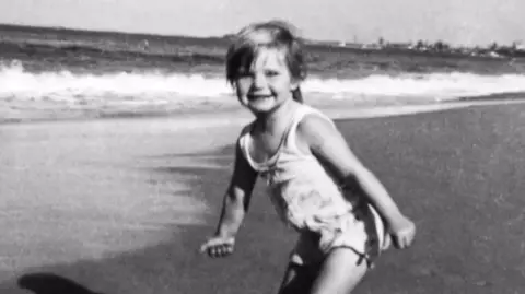 A black and white photograph showing three-year-old Cheryl Grimmer standing on a beach with waves gently lapping in the background. She has short fair hair and is wearing a white bathing costume. 