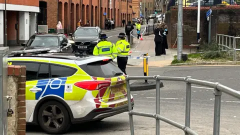 Two police officers  wearing hi-vis jackets stand near a cordon with tape up, in a street. A police car is parked in front of the cordon. There is a red building and black cars behind the officers
